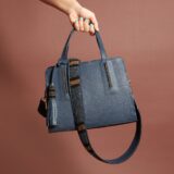 A navy blue bag with a navy, black, and brown beaded strap and tassel. A hand with blue-green nail polish is holding the bag from above.