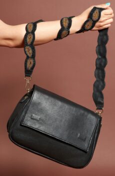 A black leather handbag with a front flap and a beaded brown and black shoulder strap. A light skinned arm is holding the bag with the strap wrapped around the forearm a few times.