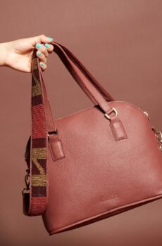A dark red leather handbag with a zippered top, handles and a beaded brown, black, and red shoulder strap.