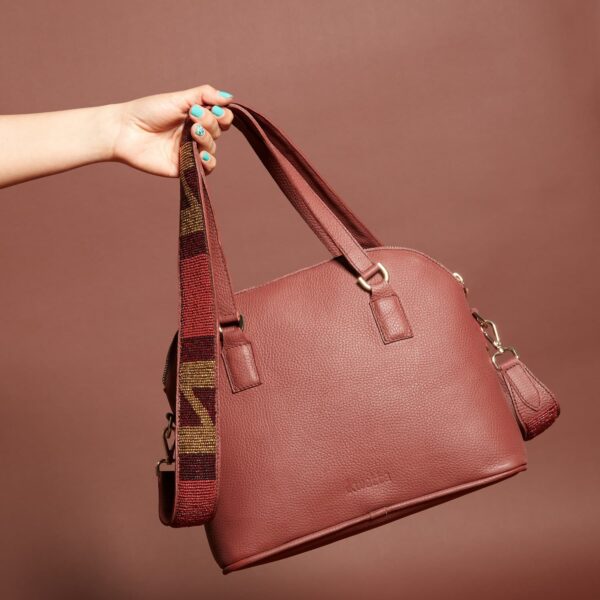 A dark red leather handbag with a zippered top, handles and a beaded brown, black, and red shoulder strap.