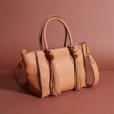 a warm tan leather bag with handles, a strap, and two tassels